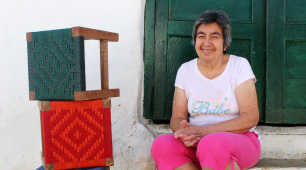 Foto de María Patrocinio Pimiento, sentada en el vestíbulo de una puerta acompañada por tres artesanías tejidas en su taller AreAgua