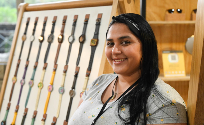 Fotografía de Melany Bolívar sonriendo y al fondo, se ven los relojes artesanales que elabora en su taller